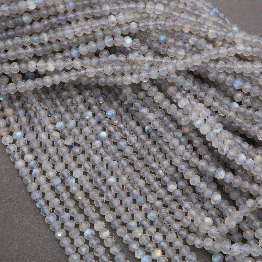 Group of labeled strands of faceted labradorite beads tied with blue strings, showing blue and gold flashes across the gray beads.