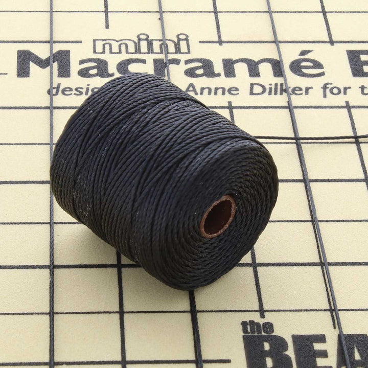 Angled view of black S-Lon TEX 210 nylon bead cord wound around a spool on a gridded macrame work surface.