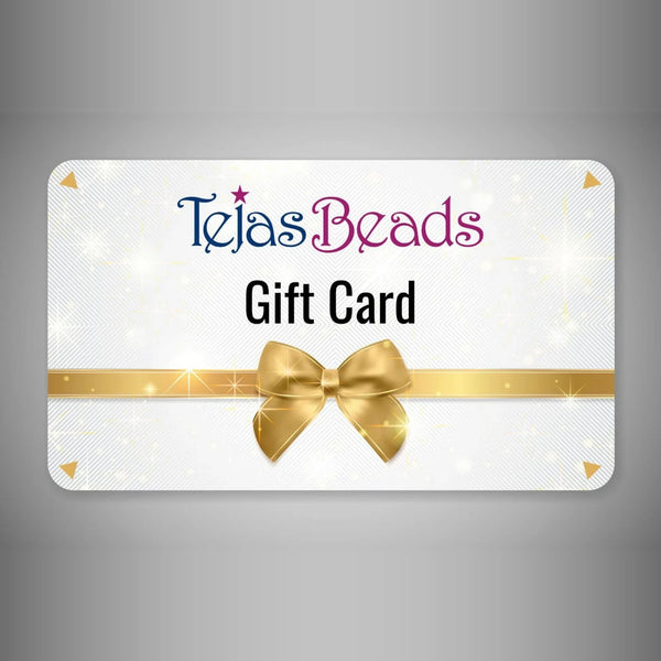 A rectangular gift card with rounded corners on a gray gradient background. The card is white with a subtle glittery texture, featuring the text 'Tejas Beads' in blue and pink, 'Gift Card' in black, a horizontal gold ribbon, and a large gold bow in the center.
