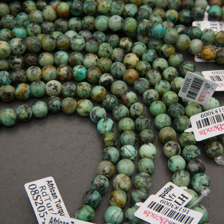 Multiple strands of round turquoise-green stone beads with dark and yellowish marbling, laid out on a gray surface with product tags visible.