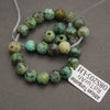A beaded strand of African turquoise beads with natural green, blue-green, and brown mottled patterns, shown on a gray background with a white product tag.