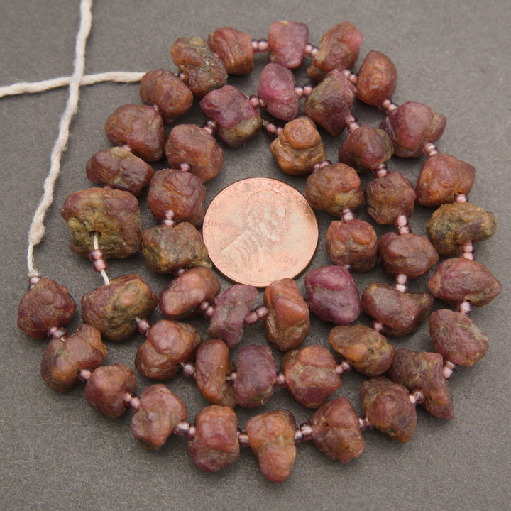 A strand of rough, polished reddish-brown and pinkish-purple stone beads strung on a white cord, displayed in a circular arrangement with a US quarter coin in the center for scale, on a dark gray textured background.
