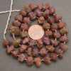 A strand of rough, polished reddish-brown and pinkish-purple stone beads strung on a white cord, displayed in a circular arrangement with a US quarter coin in the center for scale, on a dark gray textured background.