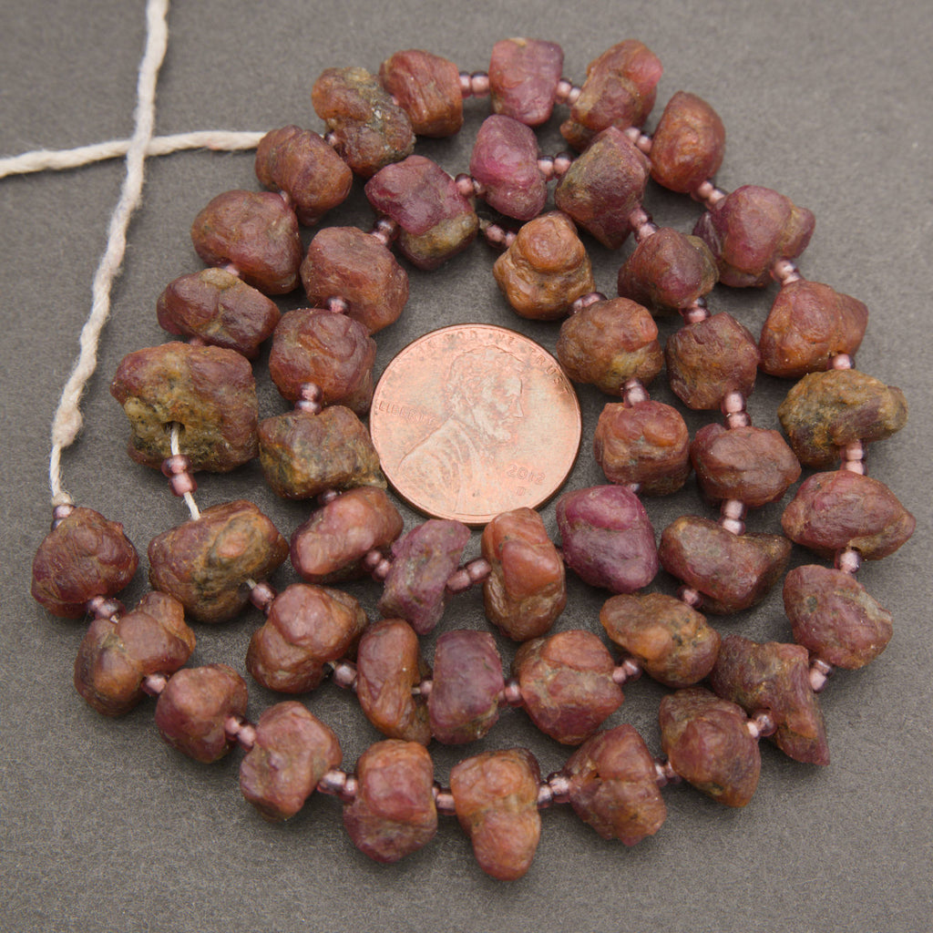 A strand of rough, polished reddish-brown and pinkish-purple stone beads strung on a white cord, displayed in a circular arrangement with a US quarter coin in the center for scale, on a dark gray textured background.