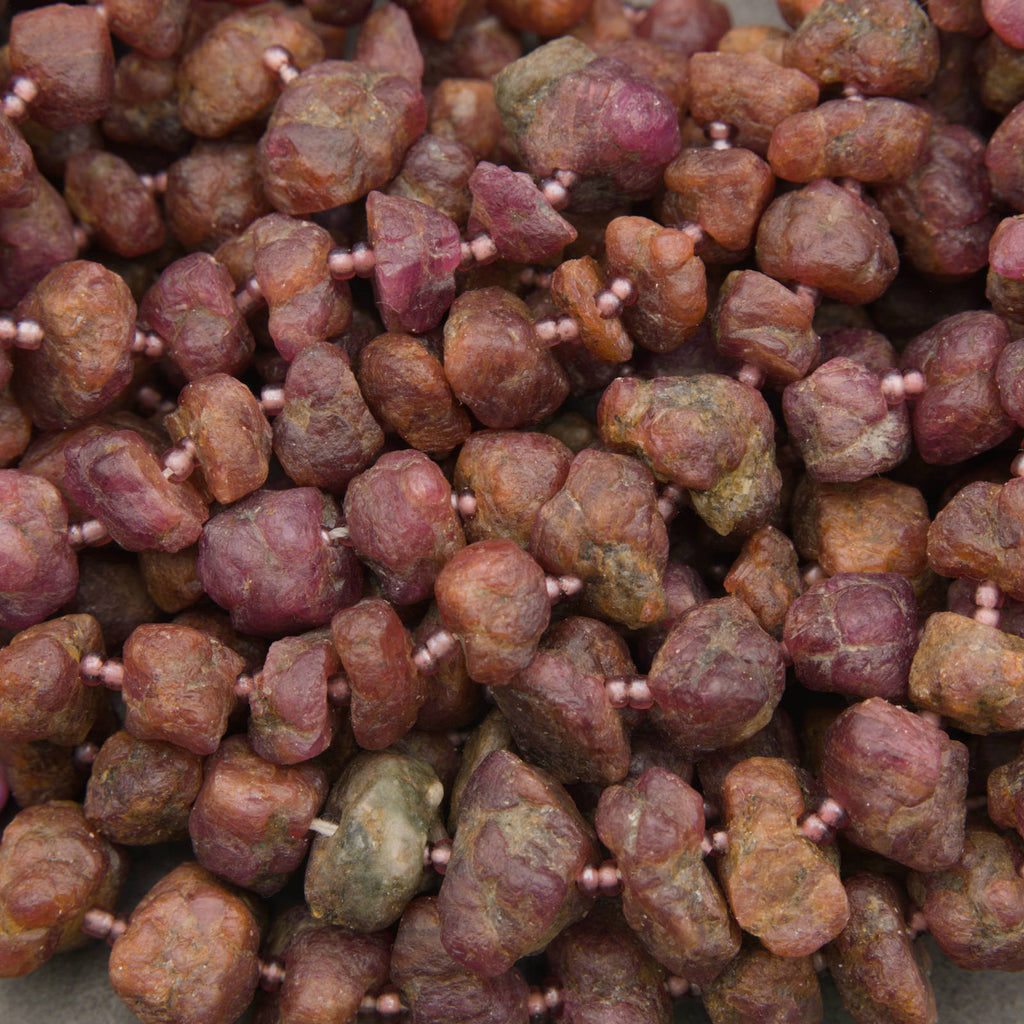 A close-up pile of rough, irregular gemstone beads strung on thin pink cords. The beads have a mix of earthy reddish-brown, dusty rose, and olive-green tones with a natural, unpolished texture.