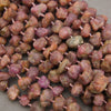 A close-up of raw, rough stone beads strung on a cord, displaying natural earthy colors of brown, reddish-brown, and purple.