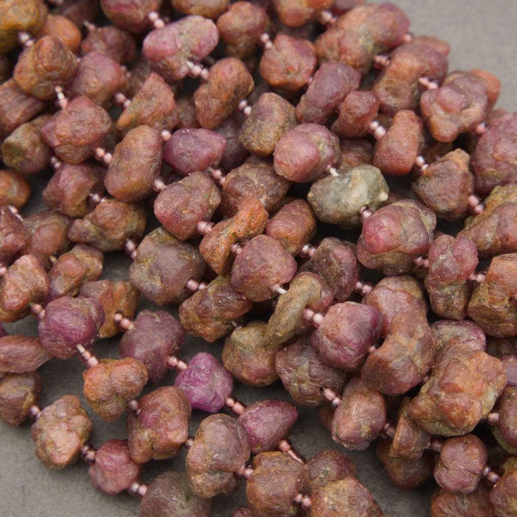 A close-up of raw, rough stone beads strung on a cord, displaying natural earthy colors of brown, reddish-brown, and purple.