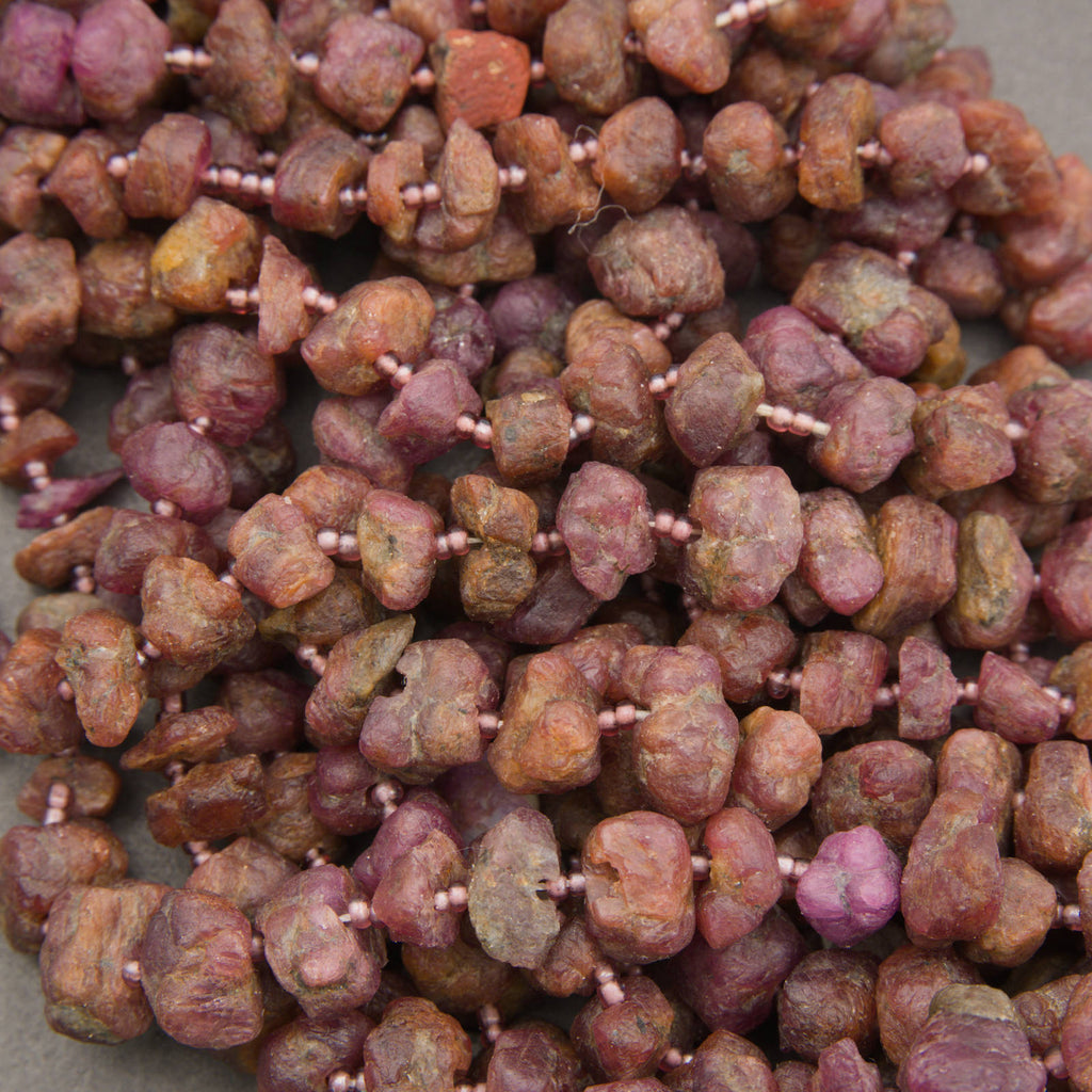 A close-up pile of rough, irregularly shaped beads strung on a cord, showing a mix of earthy brown, terracotta, and dusty rose/pink tones.