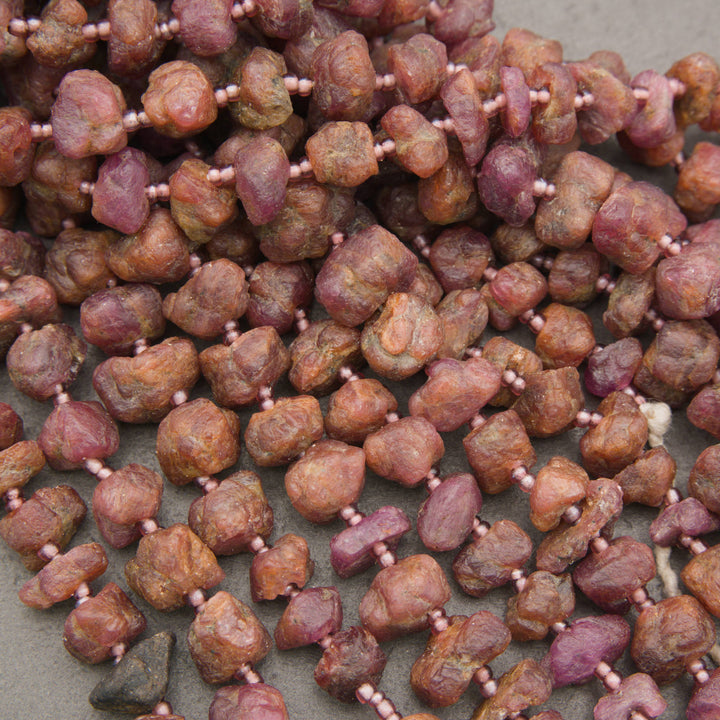 A close-up of rough, unpolished gemstone beads strung on a cord, showing natural earthy tones of brown, reddish-brown, and pinkish-purple, separated by tiny pink spacer beads.
