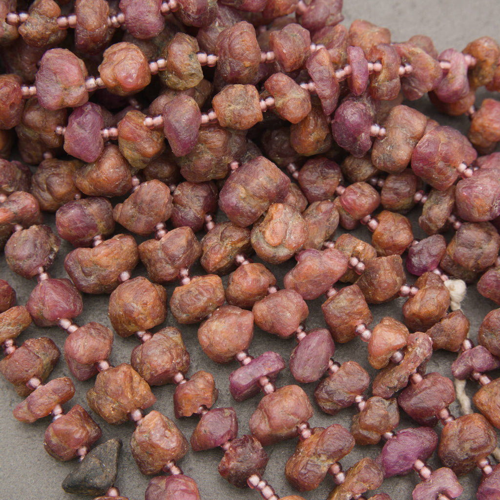 A close-up of rough, unpolished gemstone beads strung on a cord, showing natural earthy tones of brown, reddish-brown, and pinkish-purple, separated by tiny pink spacer beads.