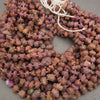 Chunky natural stone beads in earthy reddish-brown and purple tones, strung on white cord and arranged in a pile on a neutral gray surface.