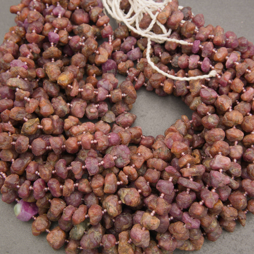 Chunky natural stone beads in earthy reddish-brown and purple tones, strung on white cord and arranged in a pile on a neutral gray surface.