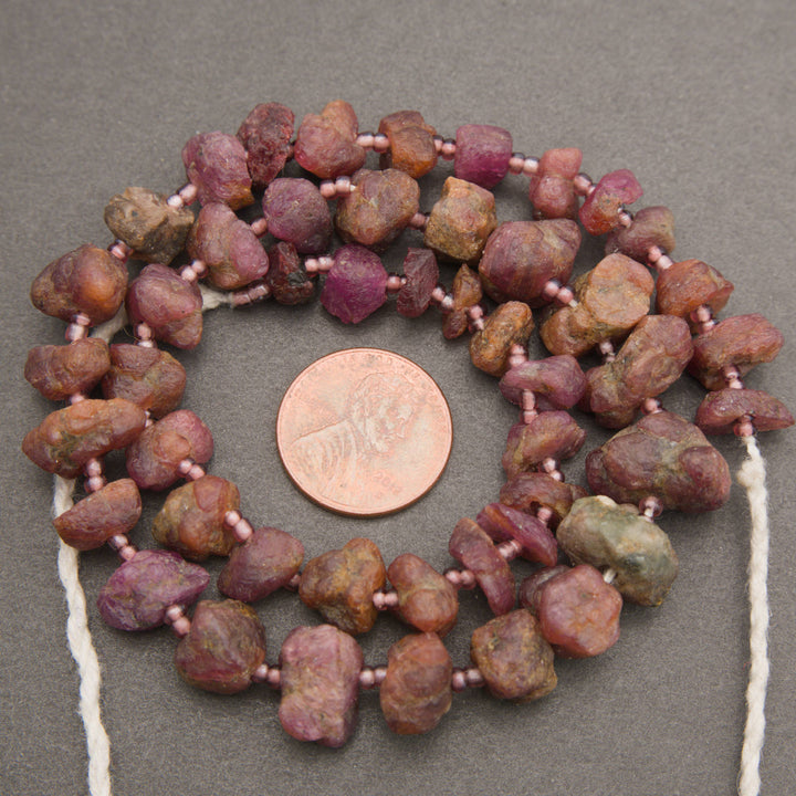A strand of rough, unpolished gemstone beads in earthy brown, pink, and purple tones, strung on white cord and displayed next to a US penny for scale on a neutral gray background.