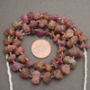 A strand of rough, unpolished gemstone beads in earthy brown, pink, and purple tones, strung on white cord and displayed next to a US penny for scale on a neutral gray background.