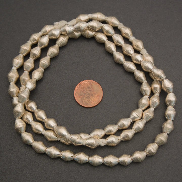 A strand of chunky, faceted metallic silver beads coiled in a circle next to a US penny for scale, photographed on a dark gray background.