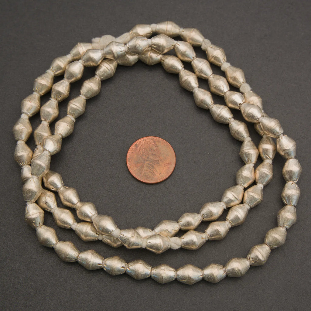 A strand of chunky, faceted metallic silver beads coiled in a circle next to a US penny for scale, photographed on a dark gray background.