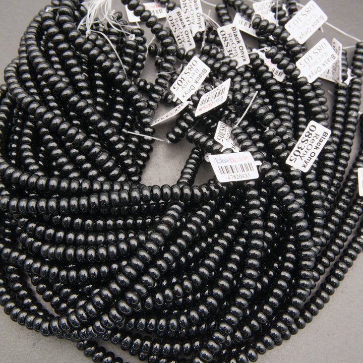 A bulk coil of glossy black seed beads with white product tags attached, photographed on a neutral gray background.
