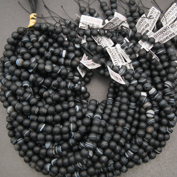 Multiple strands of matte black beaded jewelry, likely necklaces or bracelets, arranged in overlapping coils on a gray surface. Several white product tags with barcode labels are attached near the top.