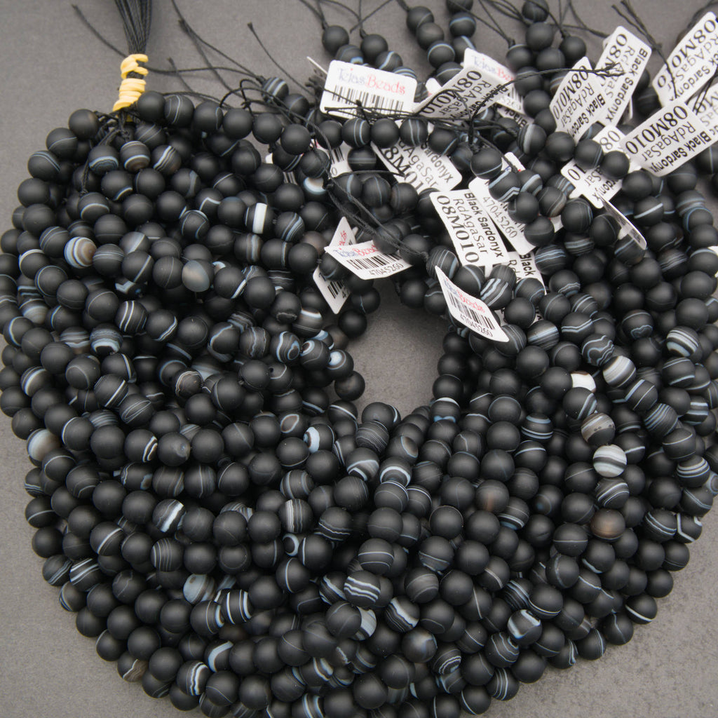 Multiple strands of matte black beaded jewelry, likely necklaces or bracelets, arranged in overlapping coils on a gray surface. Several white product tags with barcode labels are attached near the top.
