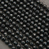 A close-up of many faceted round beads in a very dark charcoal/black color arranged in rows on a neutral gray surface.