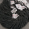 A large coil of faceted black seed beads with many white product tags attached, photographed on a neutral gray surface.