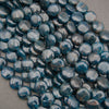 A pile of round, polished dark blue glass beads with a glossy finish and natural inclusions, resting on a neutral gray surface.