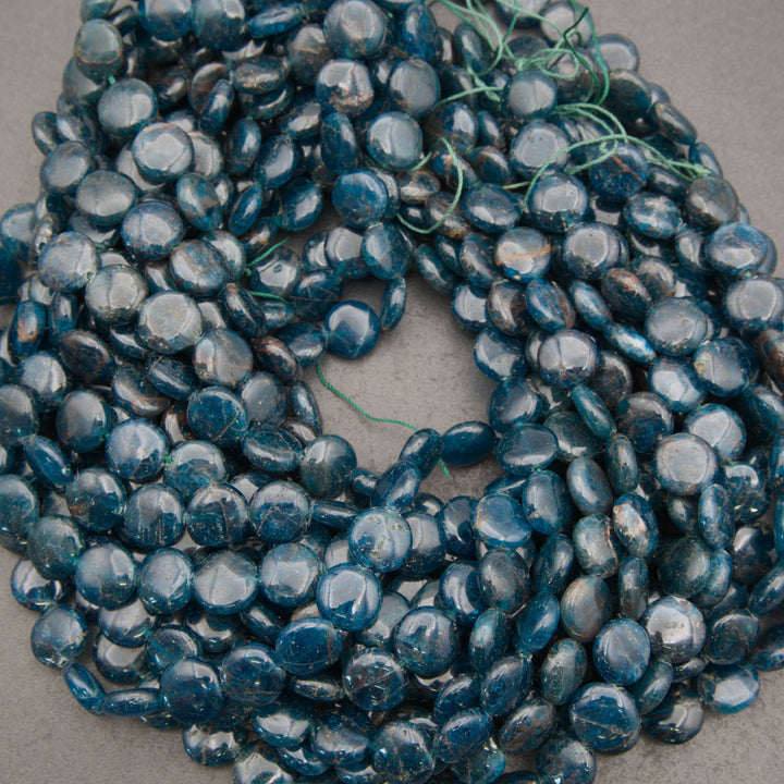 A pile of dark blue teardrop-shaped glass beads with a glossy finish, strung on thin green cords, laid on a neutral gray surface.