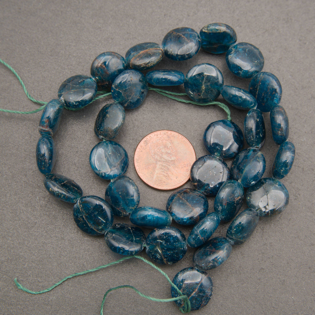 Blue polished stone beads with natural matrix inclusions, arranged in a circle around a US penny for scale on a neutral gray background.