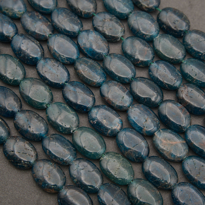 Rows of smooth oval blue apatite beads on a gray background, deep teal with rusty brown veining and natural fracture lines