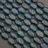 Rows of smooth oval blue apatite beads on a gray background, deep teal with rusty brown veining and natural fracture lines