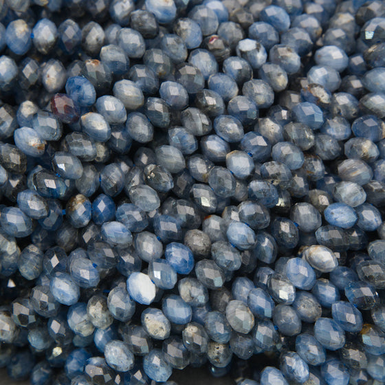 A close-up pile of faceted oval beads in a dusty slate blue color, showing their polished, reflective surfaces.