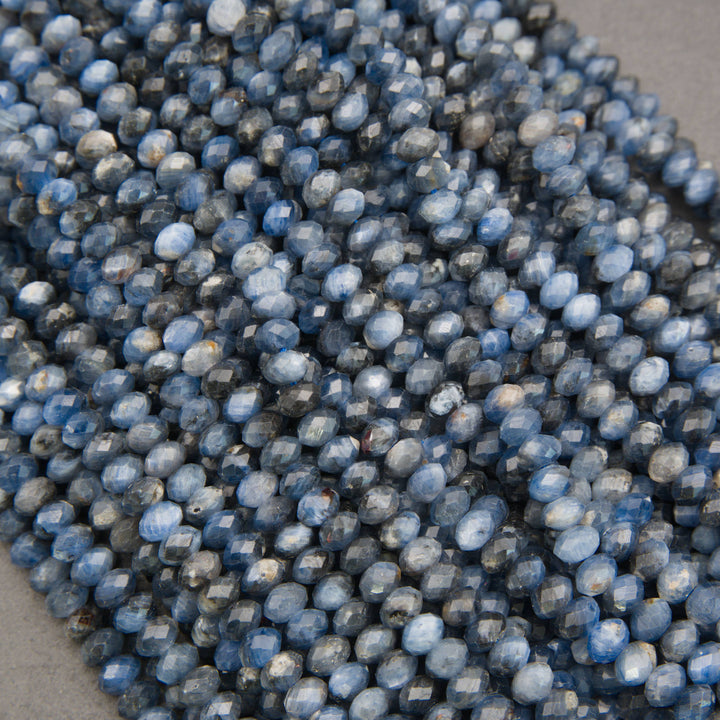 A close-up pile of faceted round beads in a dusty medium blue color with white highlights, resting on a neutral gray surface.