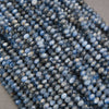 A close-up pile of faceted round beads in a dusty medium blue color with white highlights, resting on a neutral gray surface.