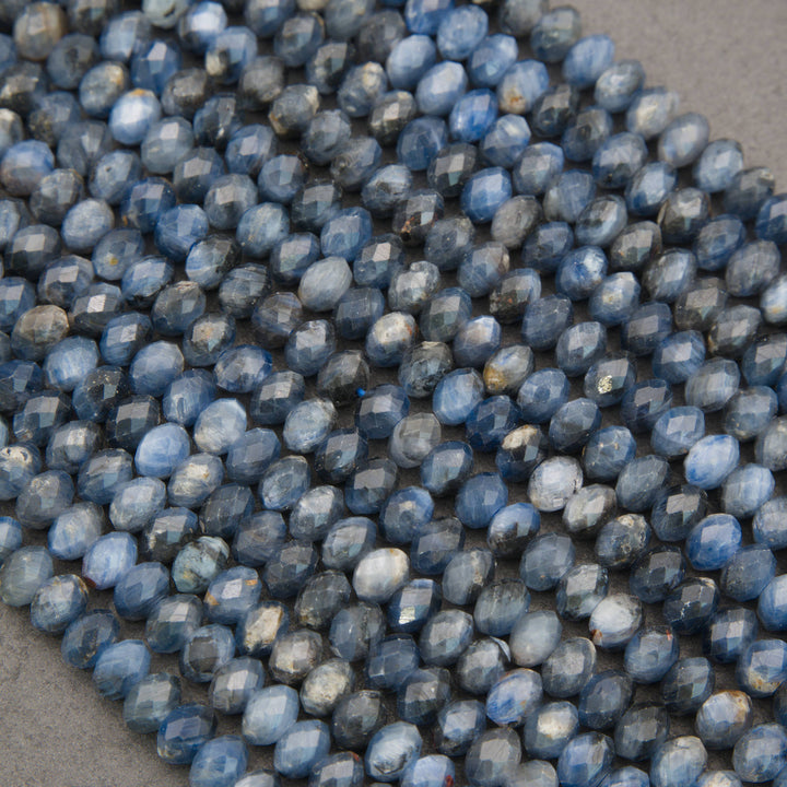 A close-up pile of faceted oval beads in a dusty slate blue color with natural mineral inclusions, resting on a neutral gray surface.