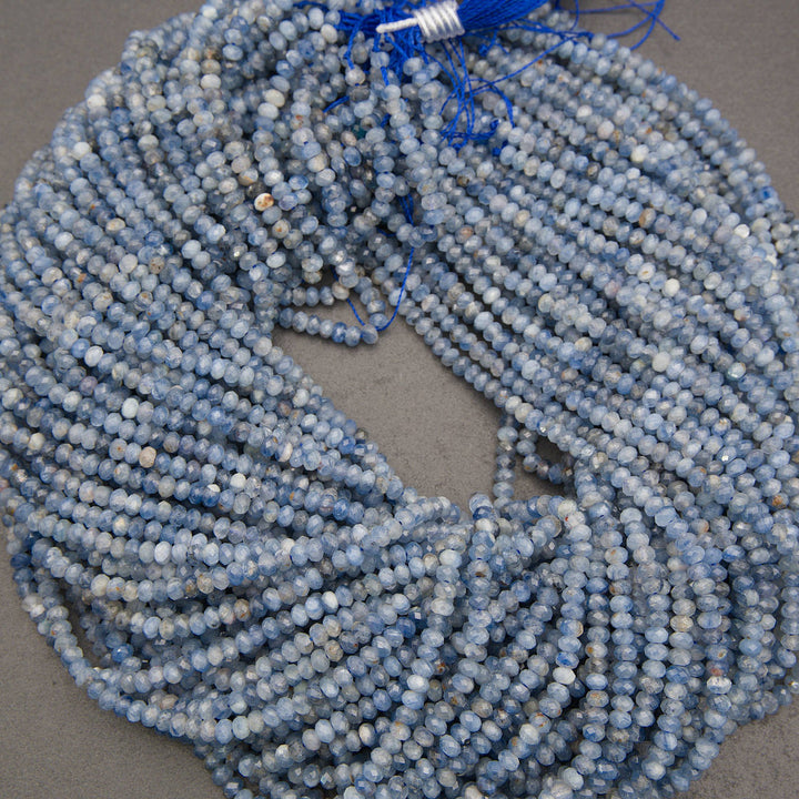 A coiled bundle of light blue and white faceted round beads with blue tassels, displayed on a neutral gray background.