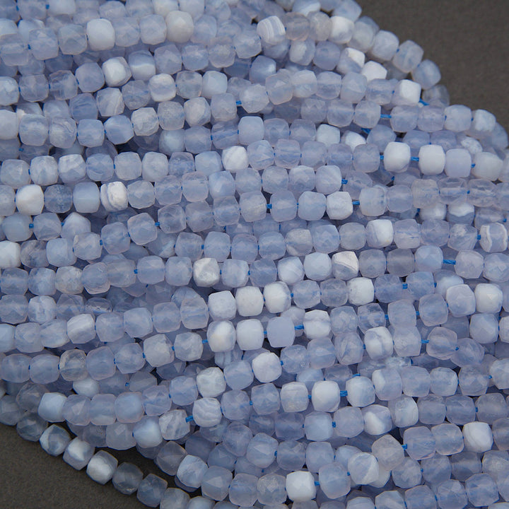 A close-up pile of small translucent pale blue beads with white cloudy inclusions, arranged on a dark gray surface.