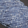 A close-up pile of small translucent pale blue beads with white cloudy inclusions, arranged on a dark gray surface.