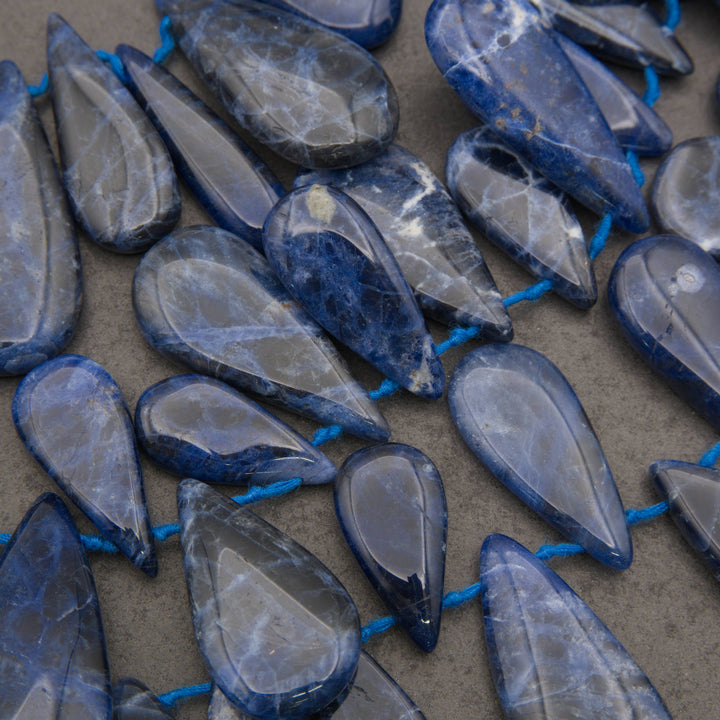 A close-up of polished blue teardrop-shaped gemstone beads strung on a cord, arranged on a neutral gray surface.