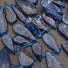 A close-up of polished blue teardrop-shaped gemstone beads strung on a cord, arranged on a neutral gray surface.