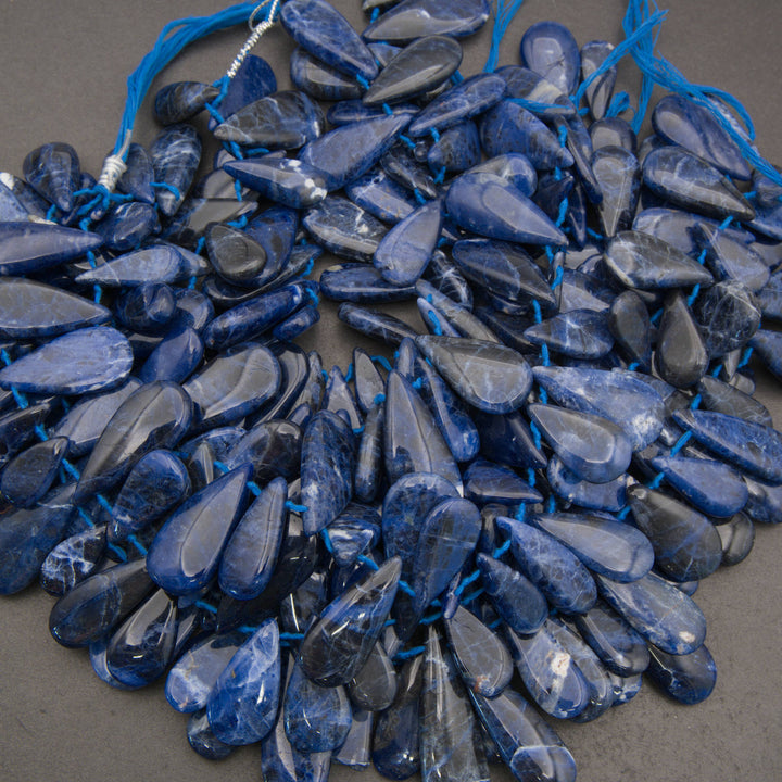 A pile of polished blue teardrop-shaped gemstone beads strung on blue cords, photographed from above on a dark gray surface.