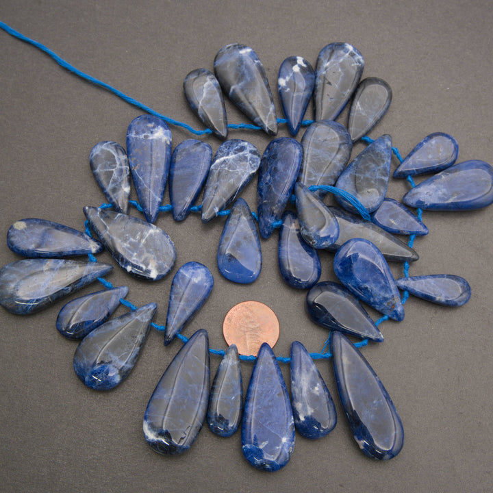 Blue sapphire teardrop beads strung on a blue cord, arranged in a circular pattern on a dark gray background with a penny for scale.