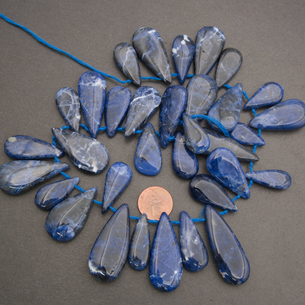 Blue sapphire teardrop beads strung on a blue cord, arranged in a circular pattern on a dark gray background with a penny for scale.