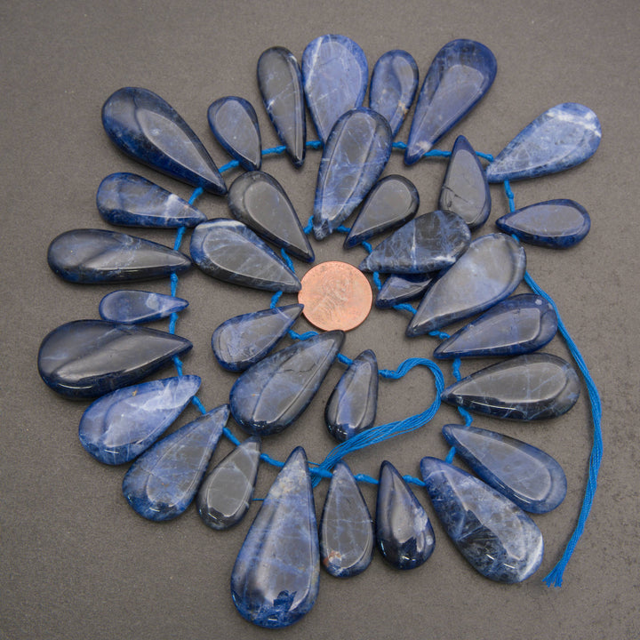 A circular arrangement of polished blue teardrop-shaped gemstone beads or pendants on a blue string, with a copper coin placed in the center for scale, photographed on a neutral gray surface.