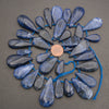 A circular arrangement of polished blue teardrop-shaped gemstone beads or pendants on a blue string, with a copper coin placed in the center for scale, photographed on a neutral gray surface.