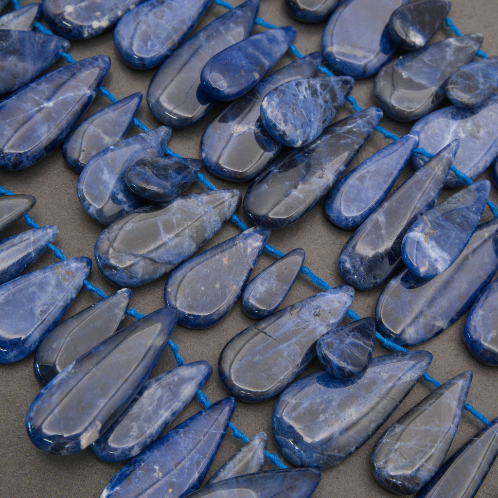 A close-up of polished blue teardrop-shaped gemstones strung along a bright blue cord, arranged in rows on a neutral surface.
