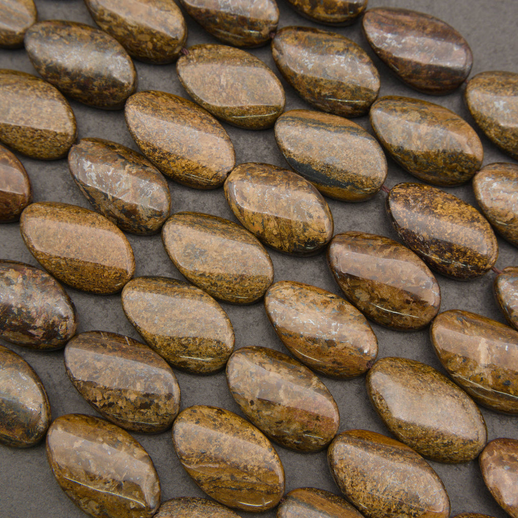 A close-up of polished oval stone beads in earthy brown, tan, and gold tones arranged diagonally on a dark gray background.