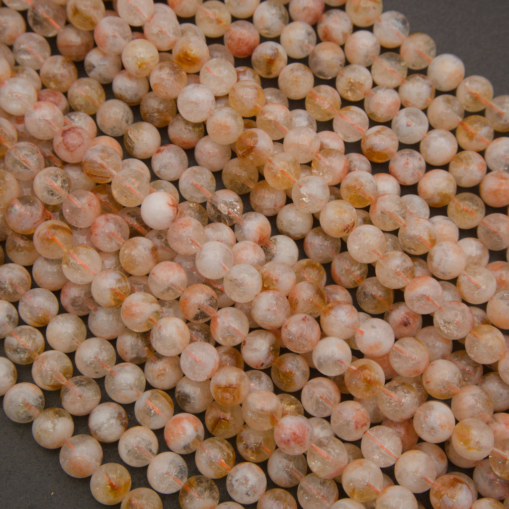 A pile of small, round, polished beads in a peachy-pink and translucent white color with subtle orange-brown inclusions, set against a dark gray background.