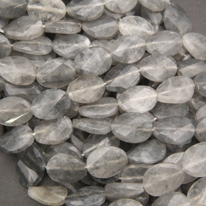 A pile of faceted oval gemstone beads in a translucent smoky gray color, showing natural inclusions and a polished finish.