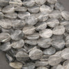 A pile of faceted oval gemstone beads in a translucent smoky gray color, showing natural inclusions and a polished finish.