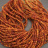 A pile of small round gemstone beads in warm orange, amber, and reddish-brown tones, with a few yellow string ties visible at the top, photographed on a gray surface.